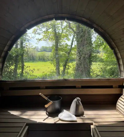 Interior view of barrel sauna showing wooden bench with sauna bucket and ladle, looking out arched window at green countryside and trees